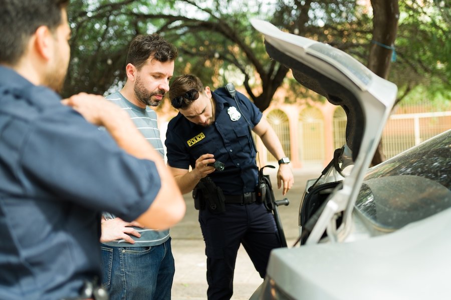Two police officers searching the trunk of the car while the driver looks on sheepishly