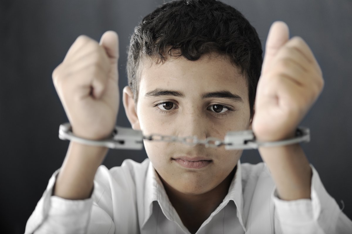 Child holding up his hands in handcuffs against a grey background