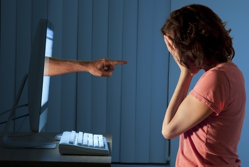 Woman with her face in her hands in front of her computer, where a menacing hand is emanating from the screen and pointing at her