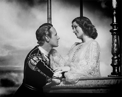 Still from the 1936 black-and-white version of Romeo & Juliet showing Leslie Howard and Norma Shearer during the balcony scene against a cloudy backdrop