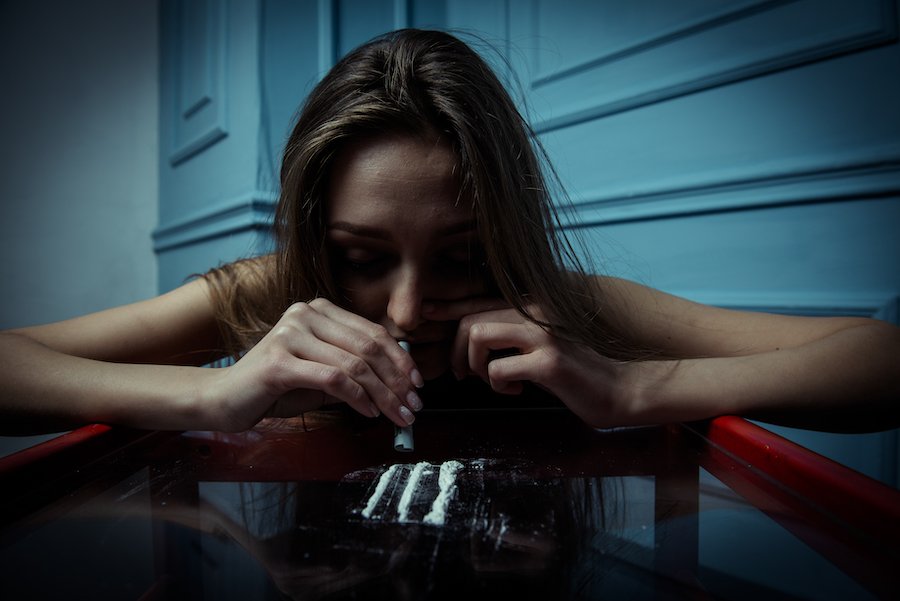 Young woman leaning over a glass table snorting three lines of cocaine against a blue background