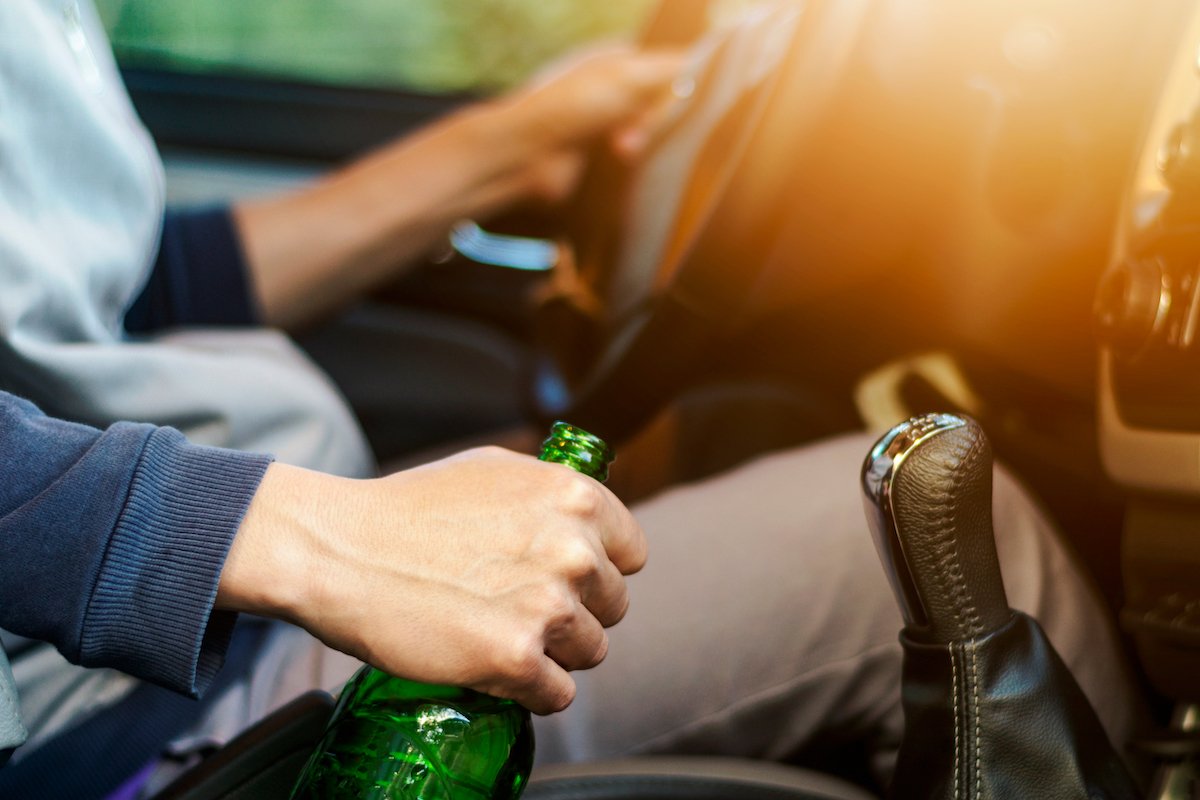 Driver holding steering wheel with one hand and a bottle of beer in the other