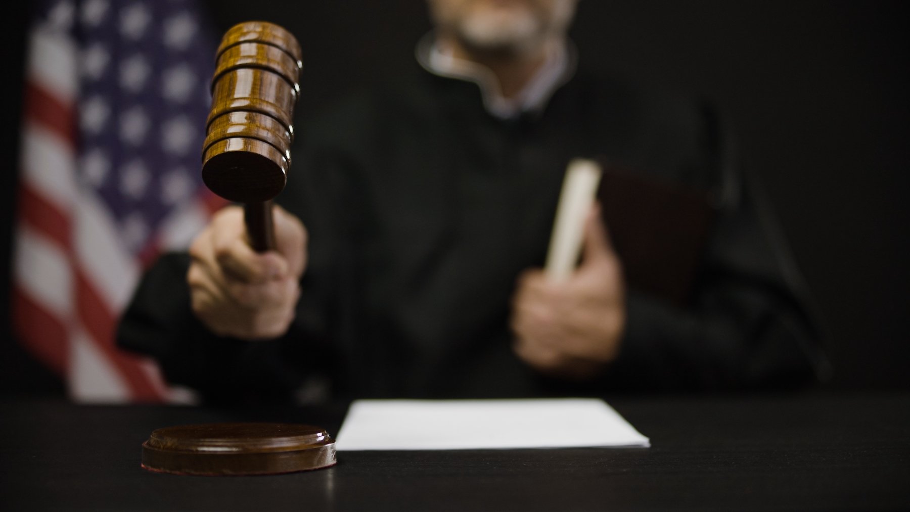Judge in black robe hammering down gavel with an American flag in the background
