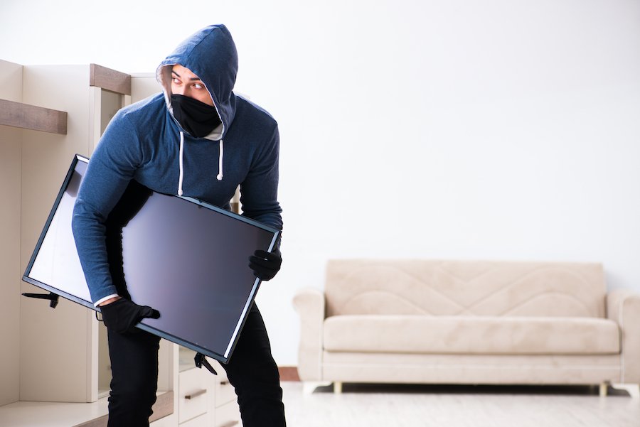 Masked and hooded adult man holding an expensive flat screen TV he is in the process of stealing in an act of grand larceny