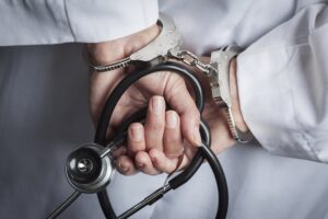 Closeup of doctor in white coat with hands in handcuffs while holding stethoscope following an NRS 686A.2815 violation