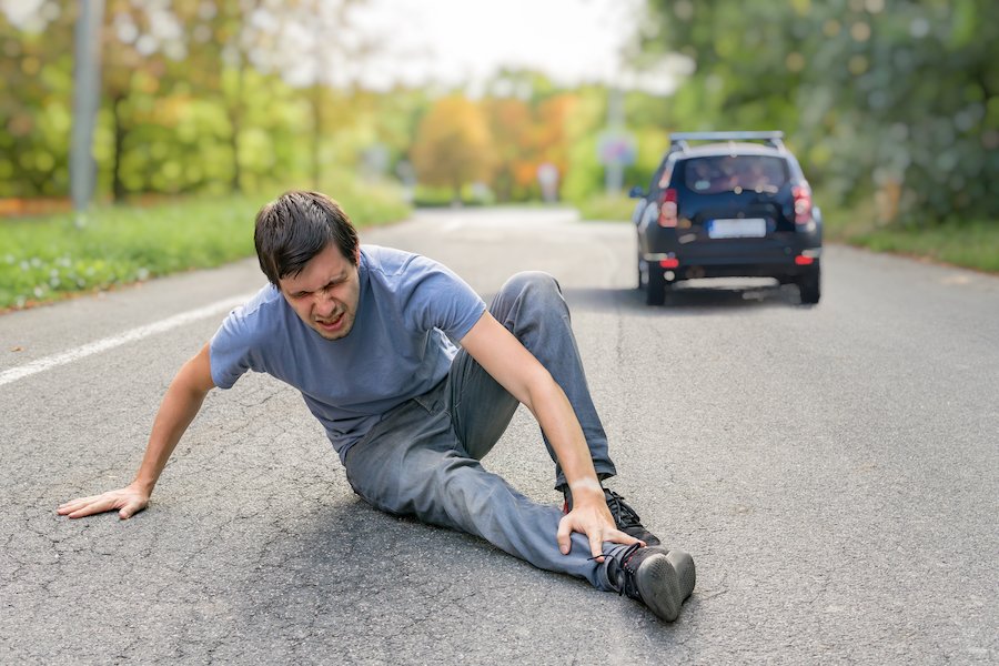 Adult pedestrian on country road clasping his injured shin after being struck by a blue car which is speeding away