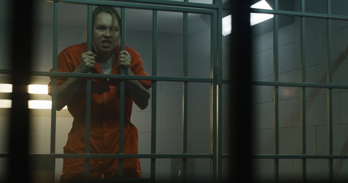 Woman inmate in an orange jumpsuit in a jail cell, and she is gripping the cell bars