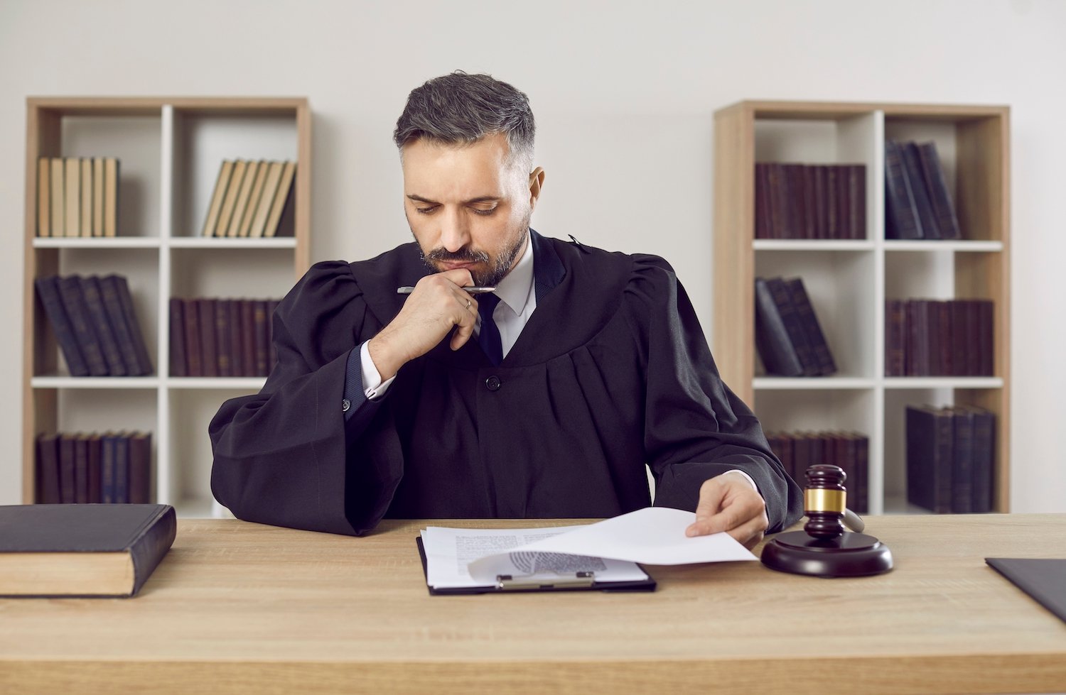 Judge in a black robe in his chambers thinking while studying papers and with a gavel next to him and bookcases in the background