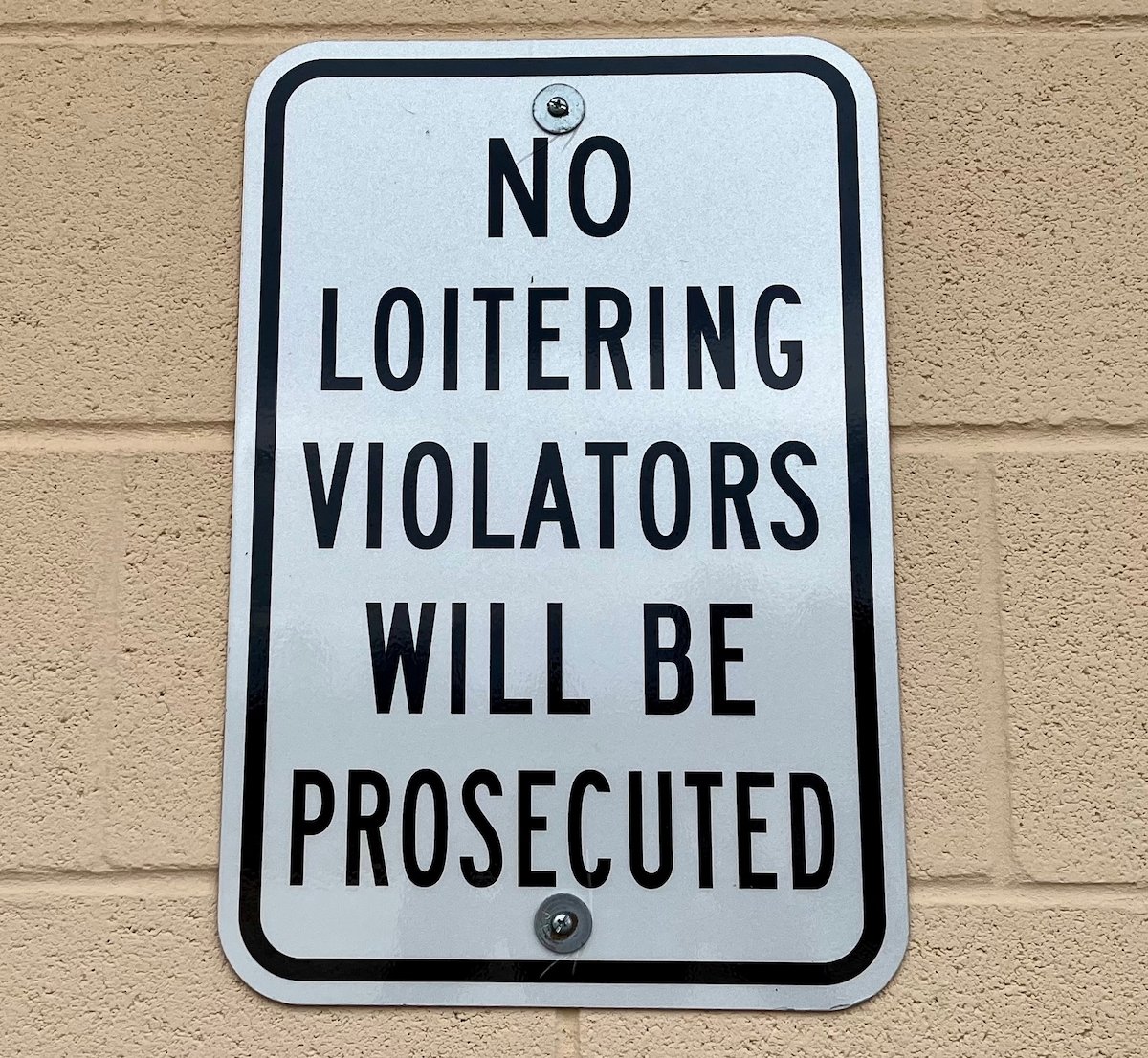 White sign with black lettering that says, "No Loitering Violators Will Be Prosecuted"