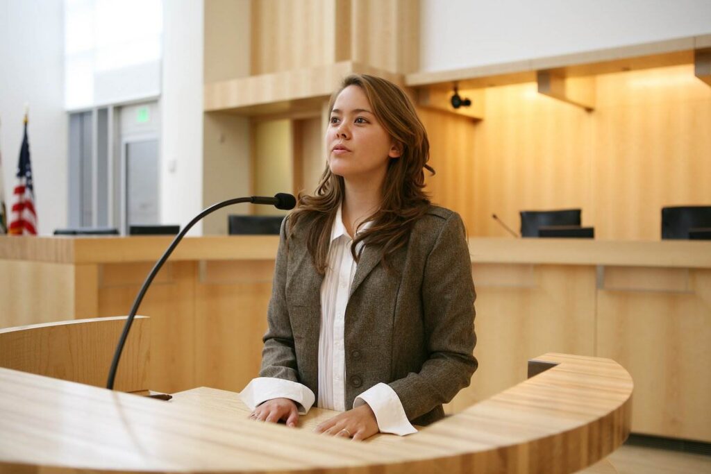 Young woman on the witness stand in a modern courtroom with an American flag in the distance and a microphone in front of her