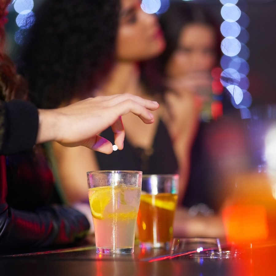 Closeup of a hand about to drop a white pill into an alcoholic drink on a bar table, against a club background showing two women oblivious to the drugging