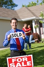 Man holding "Sold" sign in front of a "for sale" house with a happy couple in the background.