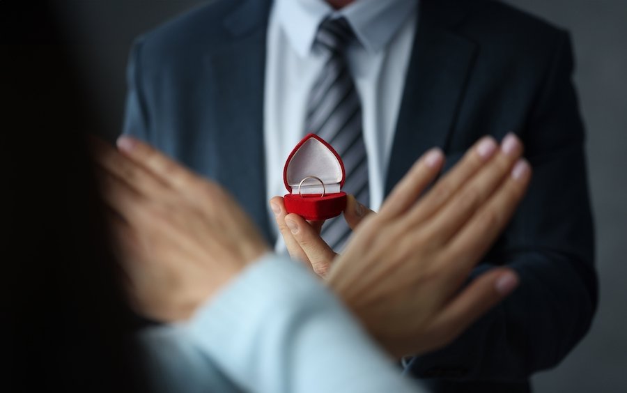 Closeup of a man presenting a wedding ring, and the recipient is holding up her hands in refusal